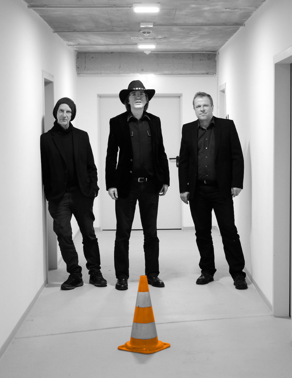 Drei Männer in schwarzer Kleidung stehen in einem weißen Flur, der strahlend hell erleuchtet ist. Der Mann in der Mitte trägt einen Cowboyhut.